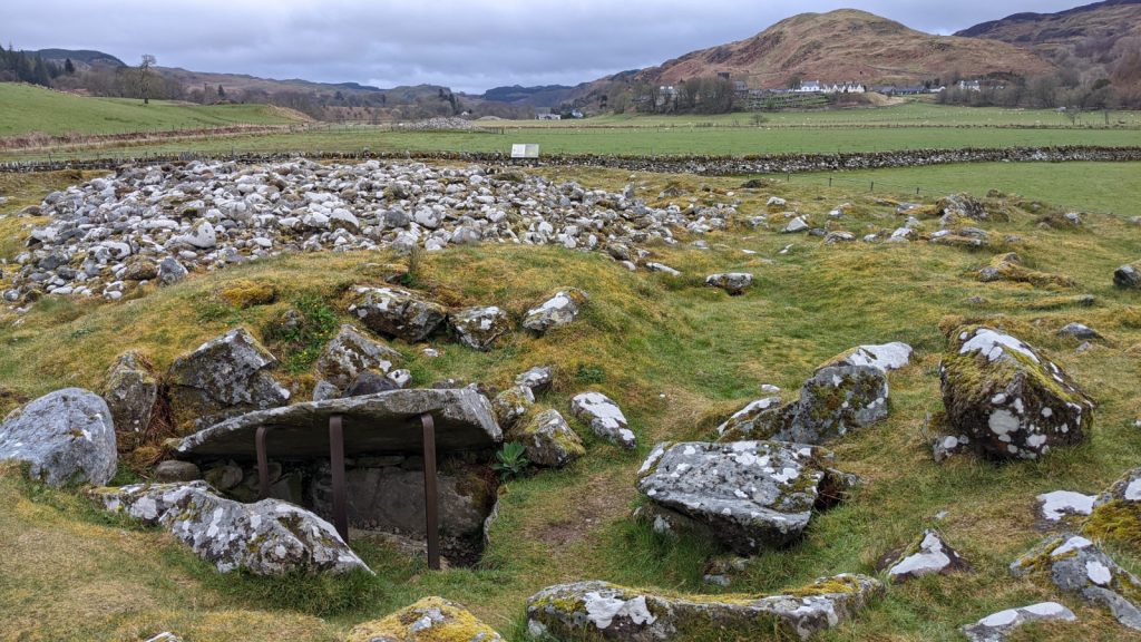 A heap of stones mark the site of an ancient cairn in a grassy field. A stone slab  is propped up, revealing a cist grave. Hills and a small village linger in the distance.