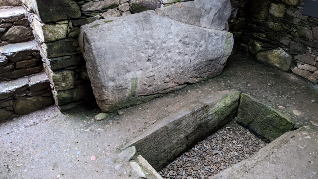 Inner chamber of a tomb, with a rectangular hole in the ground for a burial. A large rectangular stone slab rests against a wall, covered in cup marks.