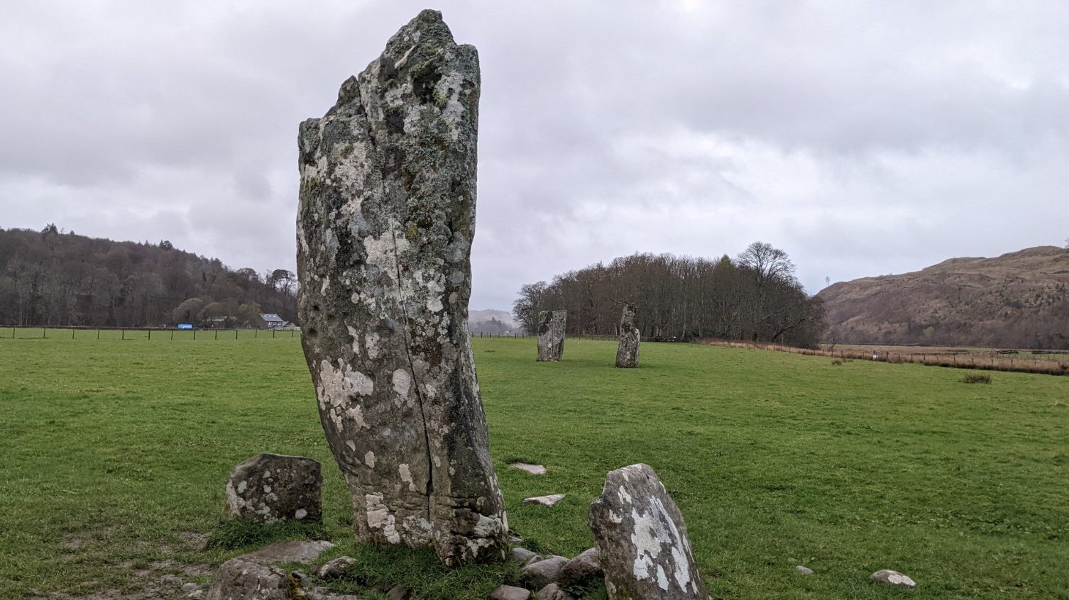 A large grey standing stone bending in an almost convex angle towards the sky stands within a grassy field, with two other standing stones beyond it positioned like hands about to clap. 