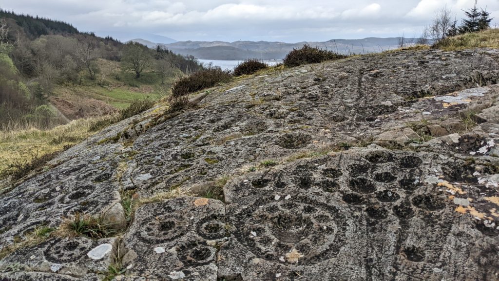 A rocky outcrop surrounded by forested hills and a loch. On the outcrop are dense patterns of rock art, some basic cups and others resembling donuts punched with many holes.