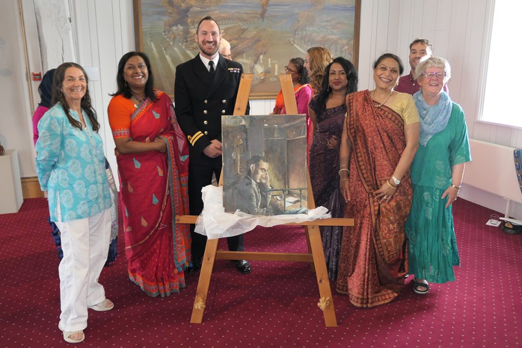 A group of South Asian and White women standing around a painting next to a dapper White man in a sailor uniform.