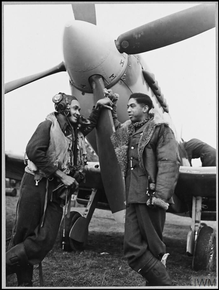 Two pilots of colour standing in front of a Spitfire's propeller, chatting and smiling.