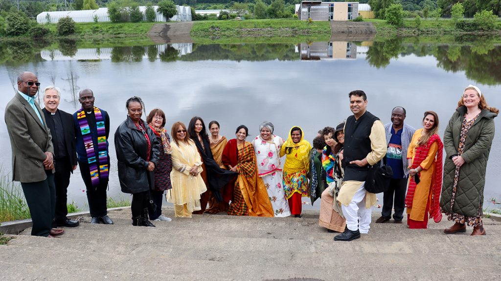 A group of Black, Brown and White people standing in a line smiling in front of a smooth lake.