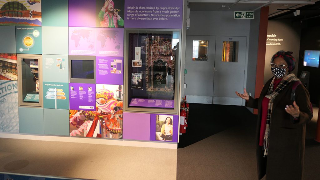 A Black woman standing in the museum in front of colourful panels exploring the diversity of Newcastle's population.