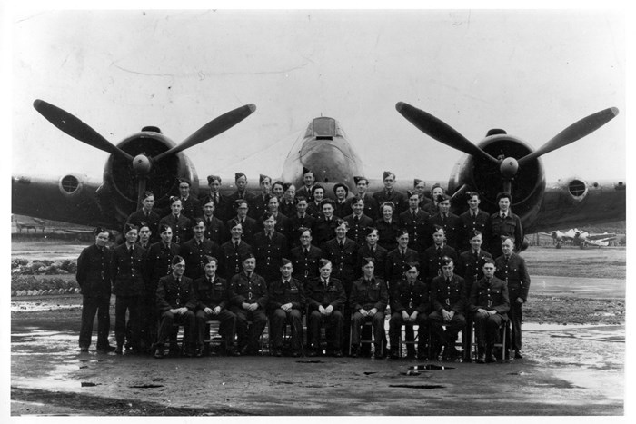 Black and white photo of four rows of aircrew in uniform stood in front of a large plane with two propellers. 