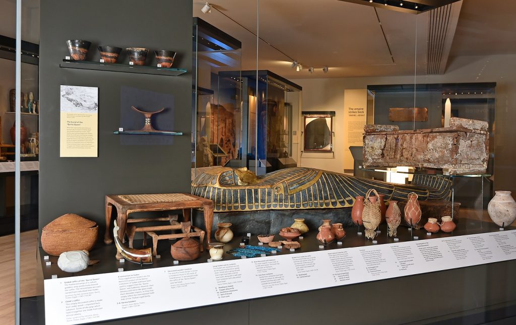 Large glass case within a softly lit museum gallery displaying many Egyptian artefacts, including a coffin case, small red pottery, a brown horn, and a small wicker stool.