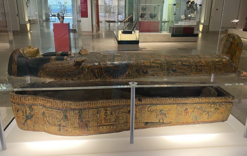 Egyptian coffin within a spacious museum gallery depicting a figure with crossed arms and braided black hair covered in hieroglyphs and dramatised scenes. 