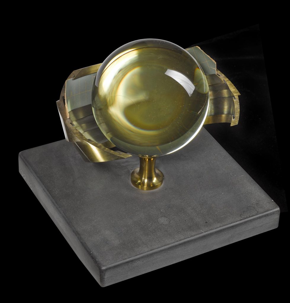 A brass curved piece of metal with a large glass sphere in front of it, mounted on slate.