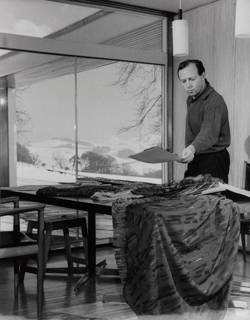The designer Bernat Klein in longsleeve shirt and black trousers examines a fabric sample on a table filled with them. A large floor to ceiling windown behind him looks out to a snowy, hilly landscape.
