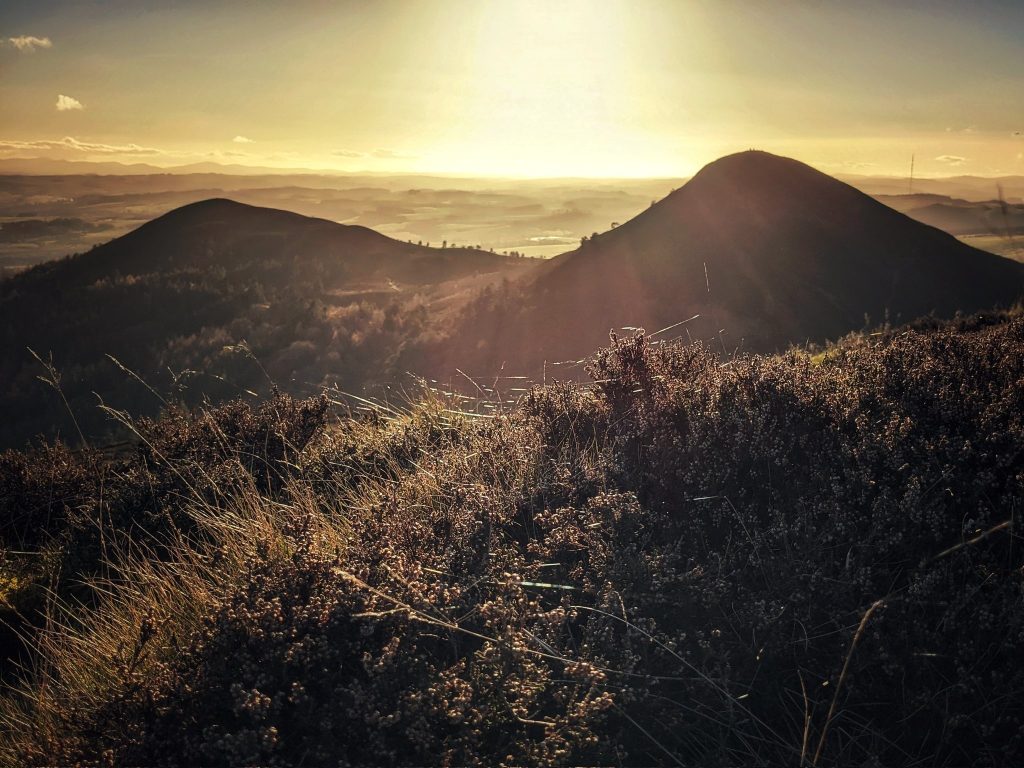 The sun sets a brilliant golden hue behind two peaks of the Eildon Hills, which are silhouetted. Rays highlight a few trees in a valley, with the foreground filled with heather covered by strands of spiders' webs. It's like you're laying in the grass watching the sun set.