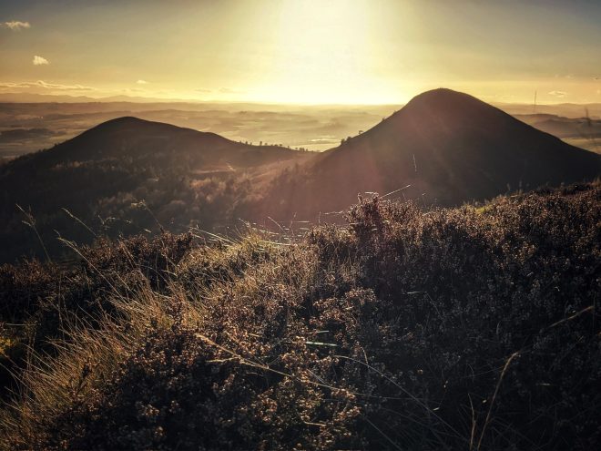 The sun sets a brilliant golden hue behind two peaks of the Eildon Hills, which are silhouetted. Rays highlight a few trees in a valley, with the foreground filled with heather covered by strands of spiders' webs. It's like you're laying in the grass watching the sun set.