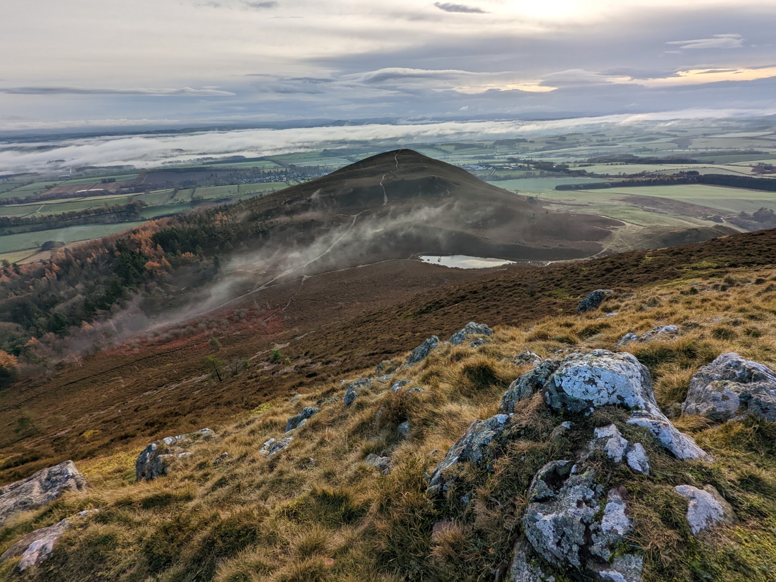 View from atop Eildon Mid Hill. Tumbled stones amid yellowed grass direct the eye to the peak of a brown hill and wisps of mist in the middle distance. Vast tracts of the Scottish Borders extends beyond and meet a wall of mist miles away.