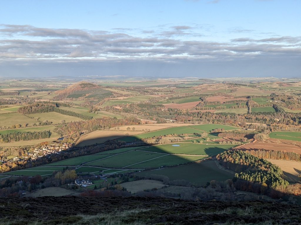 Sprawling landscape view of rolling hills, the River Tweed, and patches of forest seen from atop Eildon Hill North. The shadow of Eildon Hill North creeps over the land, covering half of it in a pyramid shape.