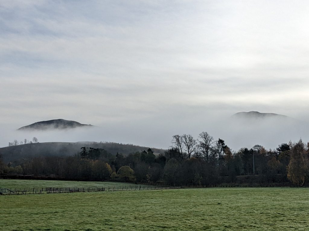 The peaks of two of the Eildon Hills barely emerge from a thick layer of mist in the distance, seen across a grassy field.