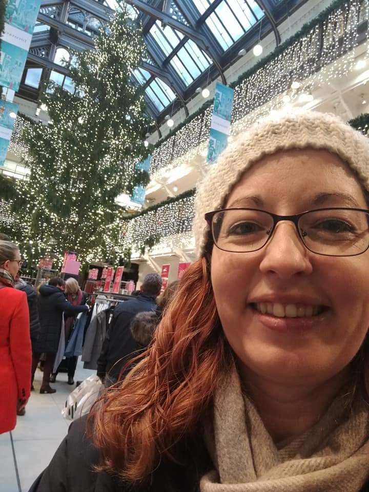 A smiling woman with red hair cascading out from under a white knitted hat poses inside the modern Jenners building. A tall Christmas tree and many Christmas lights are in the background.