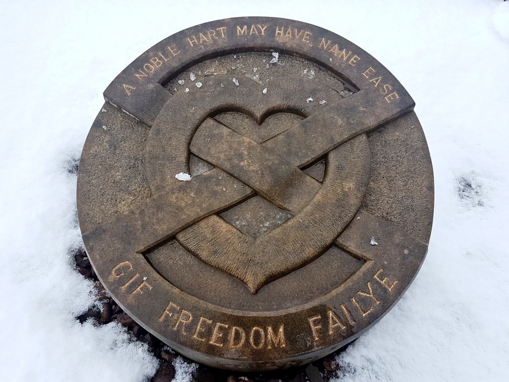 A simple stone roundel, small and very low to the ground, in the snow. A heart shape and saltire decorate it with the words 'A Noble Hart May Have Nane Ease Gif Freedom Failye'.