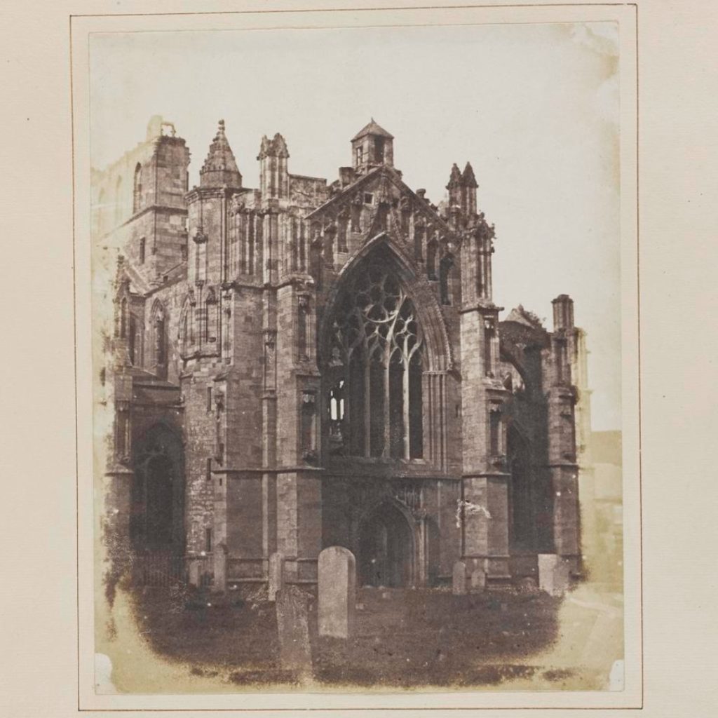 Sepia-coloured salt print, somewhat worn with time, of a closeup of a highly decorated section of Melrose Abbey. Several grave stones populate the foreground.