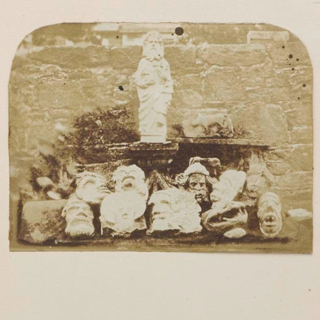 Sepia-coloured salt print showing a variety of carved heads from Melrose Abbey, arranged almost like a sports team's lineup. The heads include those of bearded people, dogs, and strange creatures.