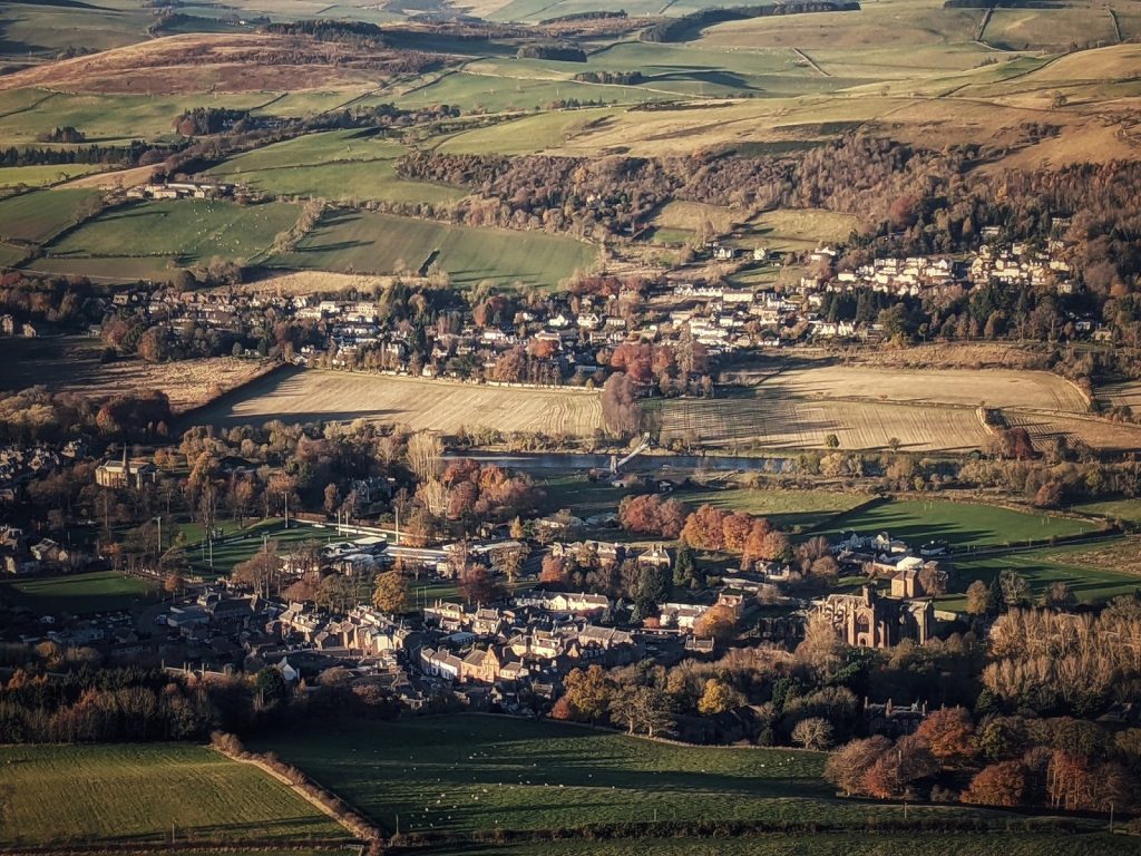 View from high in the Eildon Hills looking down on Melrose town and abbey, which look like doll's houses from so high up. The River Tweed cuts through the middle of the image, with rolling hills extending to the horizon.