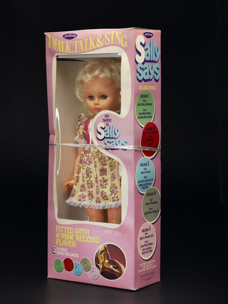 A 'Sally Says' doll, blonde with floral dress, still inside its retail box. The box boasts, 'I walk, talk & sing'.
