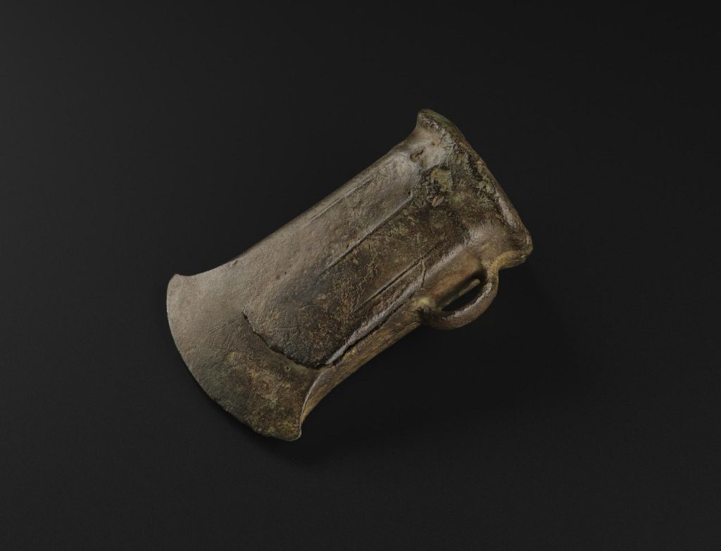 A bronze axehead, darkened with time, against a black background. The axe's edge is gently curved, and a small socket is fitted to the bottom.