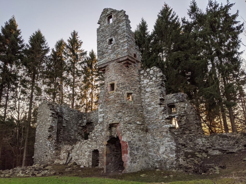 A crumbling, grey stone castle surrounded by tall conifers. The central tower stands tall with a box-like structure at the top. The roofless walls are partially collapsed, with stone rubble lying around. The main door is lined with pink stones.