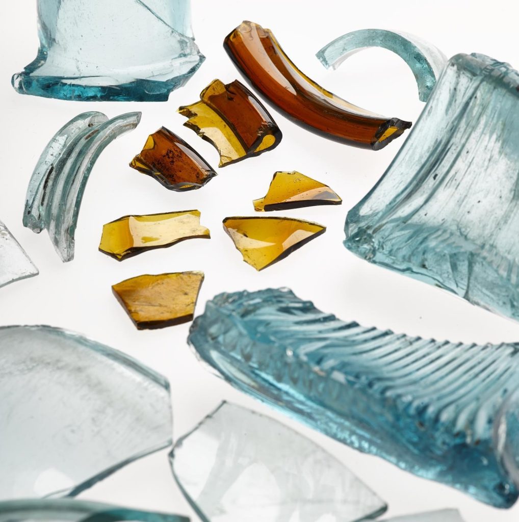 A scattering of glass fragments against a white background. Th larger fragments are translucent blue and look very modern. Smaller fragments are yellow-brown, including the rim of a vessel.