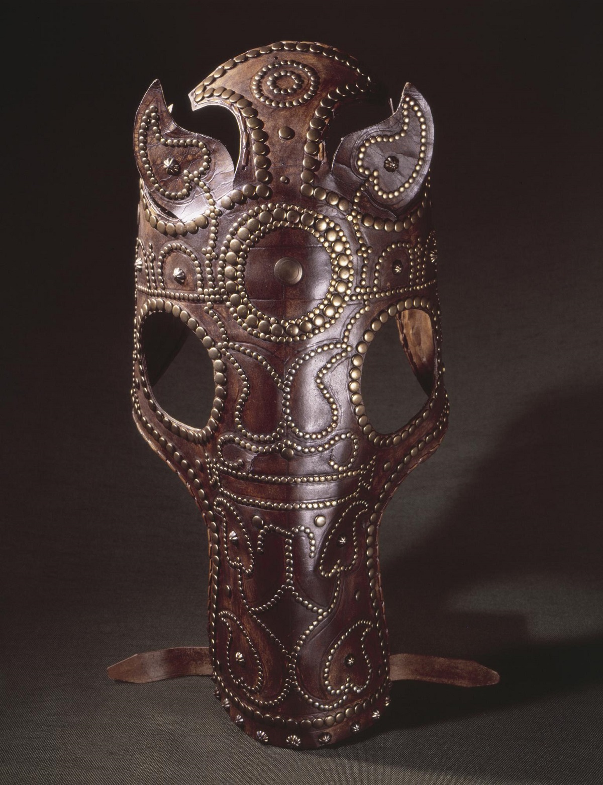 Red-brown leather chamfron or horse's face covering positioned upright on a grey background. The chamfron has eye and ear holes, with bronze studs around its edged and forming vine-like designs across the front.