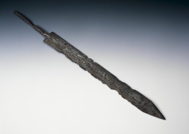 The blade and tang of a Roman sword or gladius, facing diagonally down to the right against a grey-blue background. The sword lacks a hilt and looks battered and used.