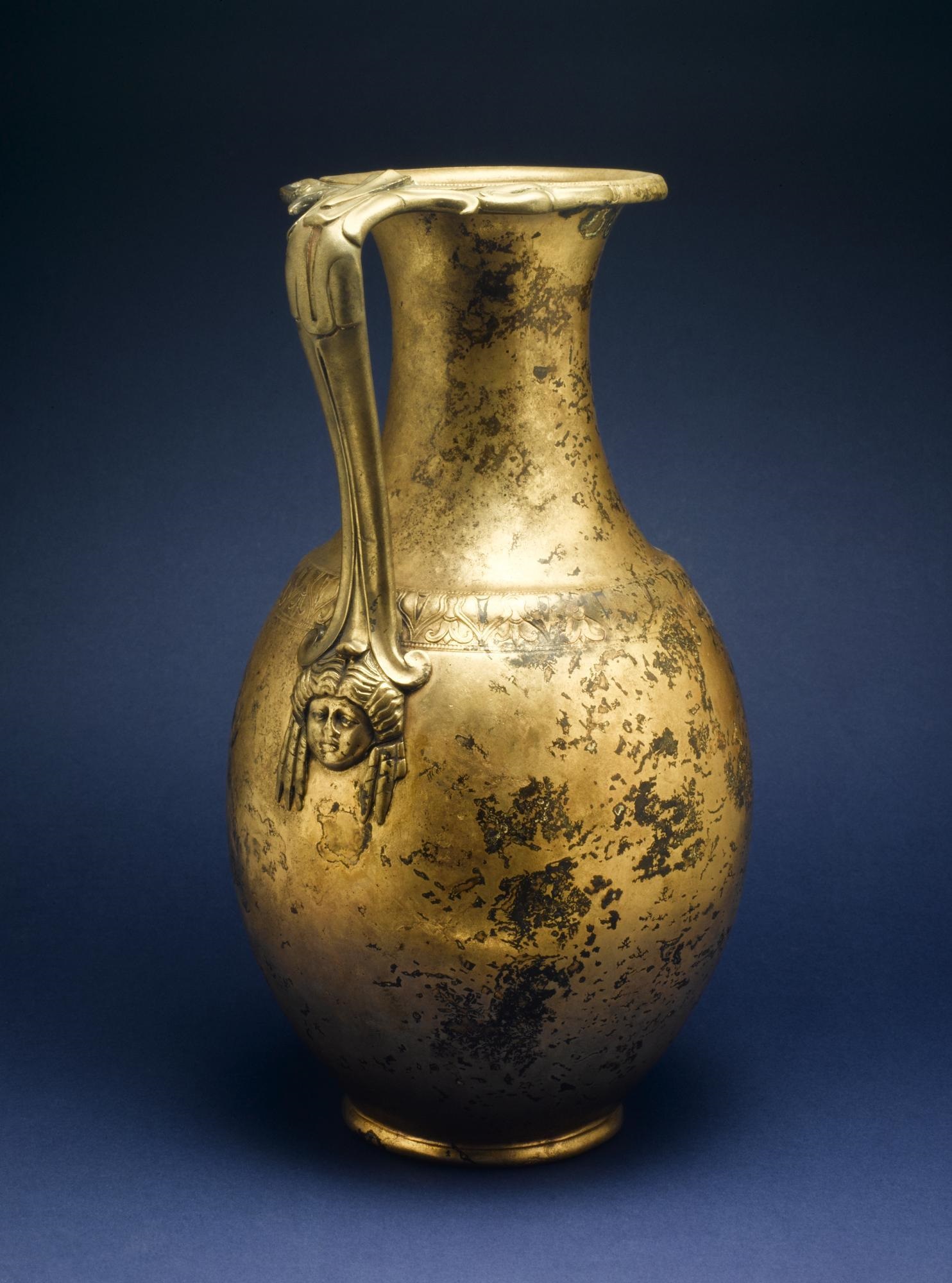 A tall, slender, and bright gold-coloured jug against a royal blue background. The jug is elegant, with a face with curly hair below the handle. Patches of darkened wear cover about one third of the surface.