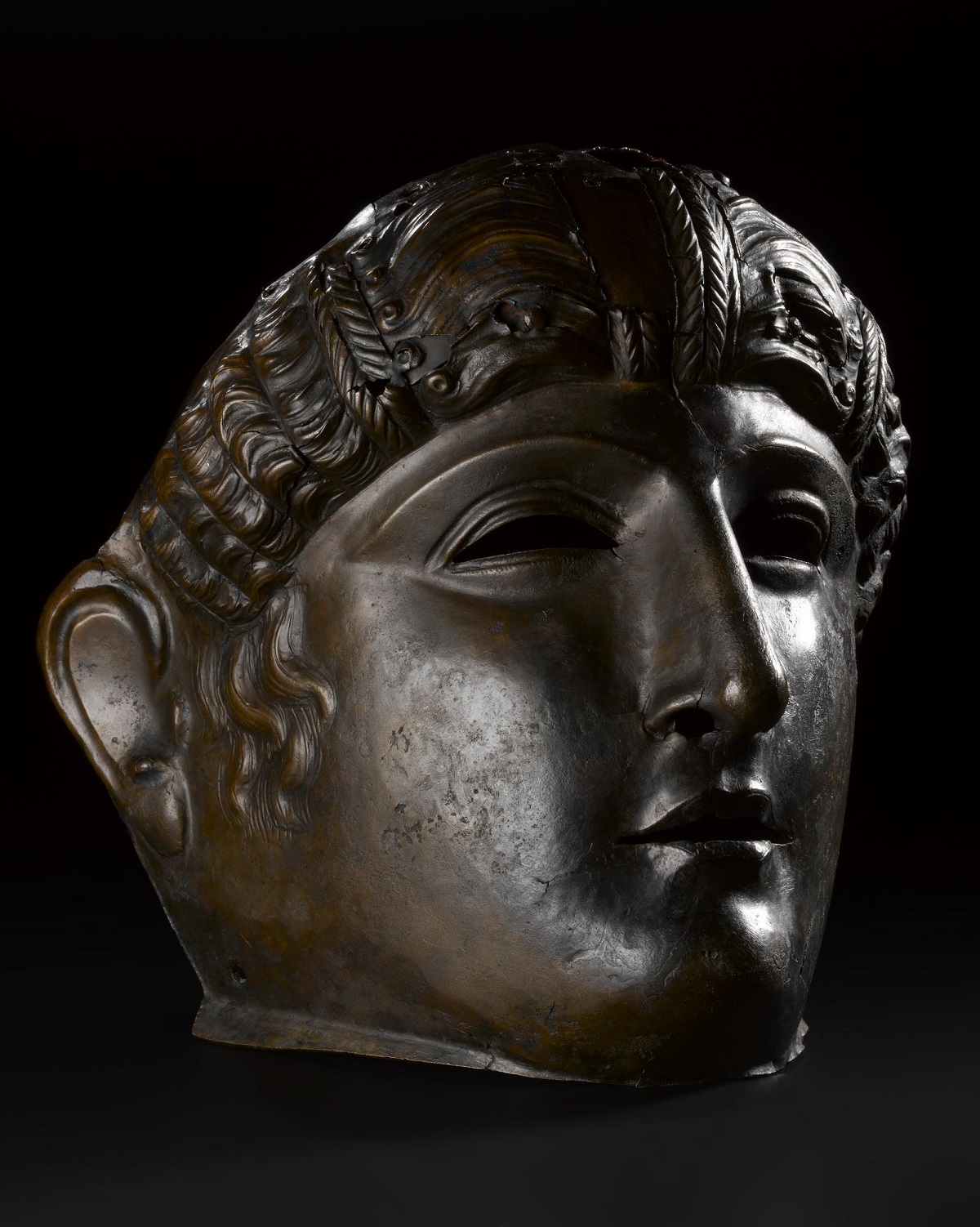 A brownish-black metal Roman parade mask in the visage of a young person with rounded cheeks, strong angular nose, and curling hair.