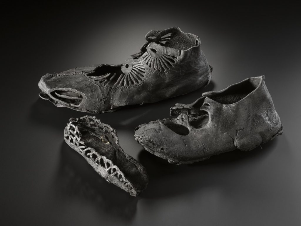 Three leather shoes in varying states of degradation on a shiny black surface. Two larger, more intact shoes with radial designs face left, and a smaller shoe's sole and sides faces right.