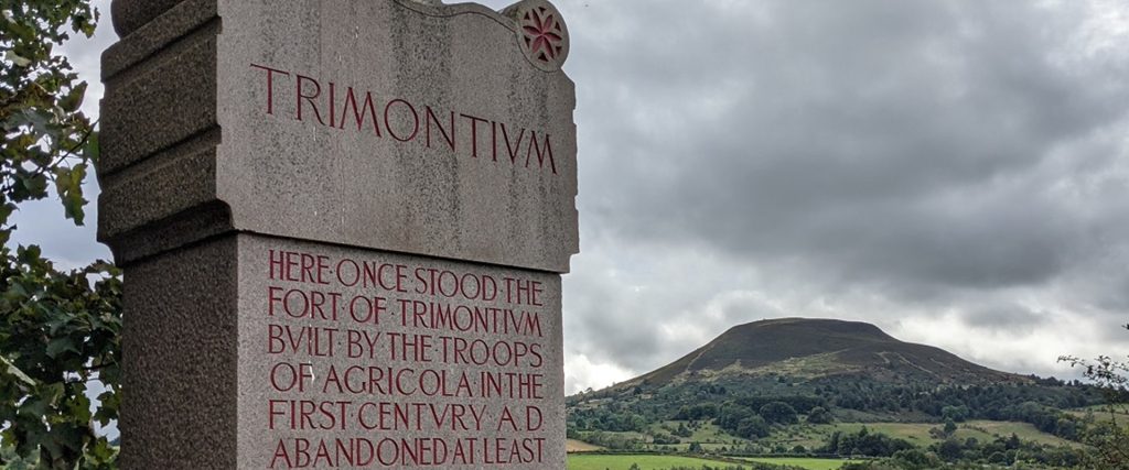 Narrow, wide view of the whaleback shape of Eildon Hill North in the distance on the right, with a modern stone altar inscribed with 'TRIMONTIUM' taking up the foreground on the left. Grey clouds loom overhead.