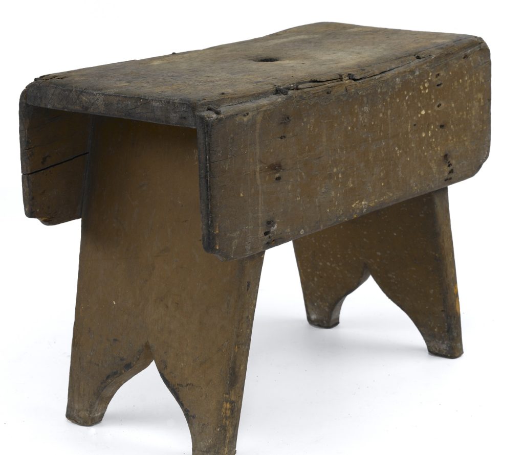 A simple, small wooden stool, dark brown in colour with plenty of signs of daily use. The top is rectangular, with two plank-like legs supporting it.