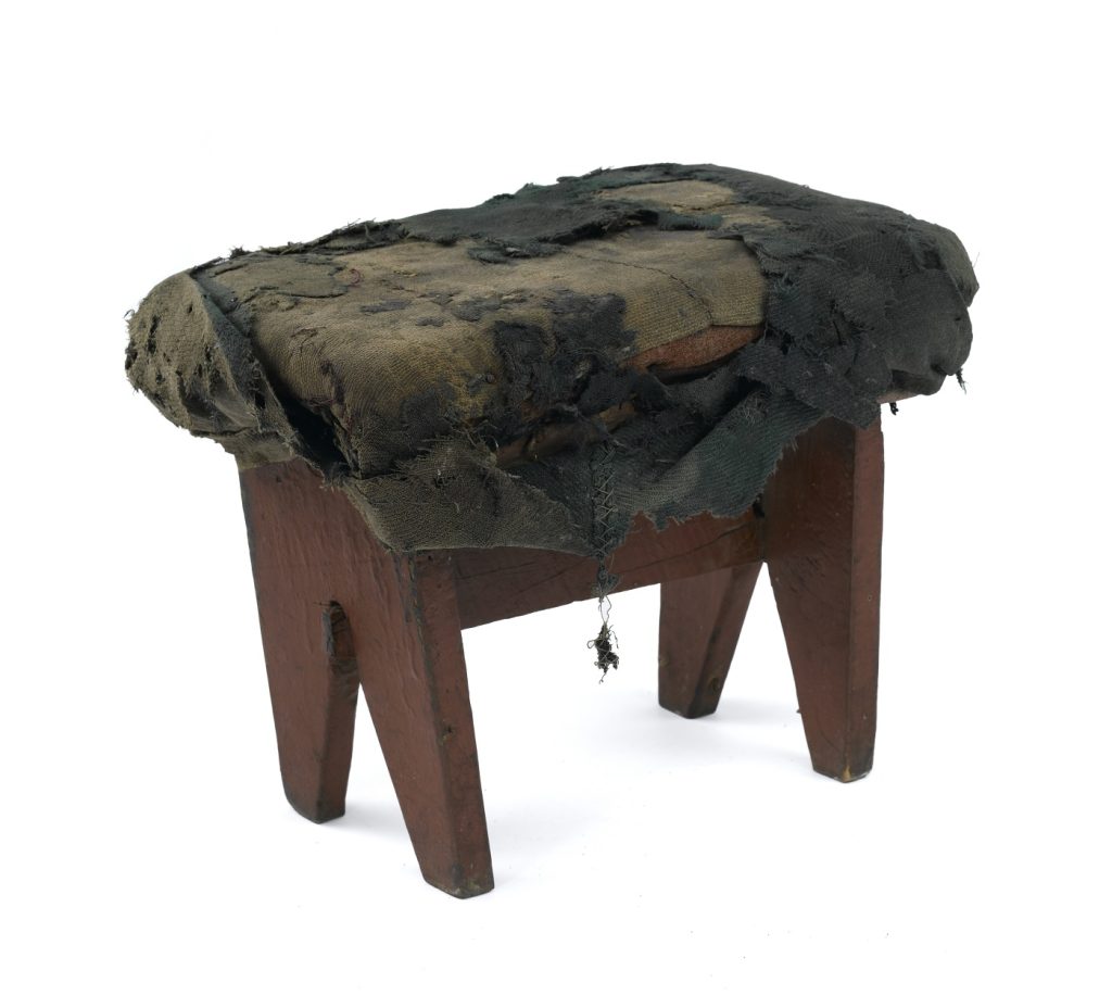 A simple, small and very worn-out wooden stool against a black background. The top is covered by a badly torn and worn green covering.