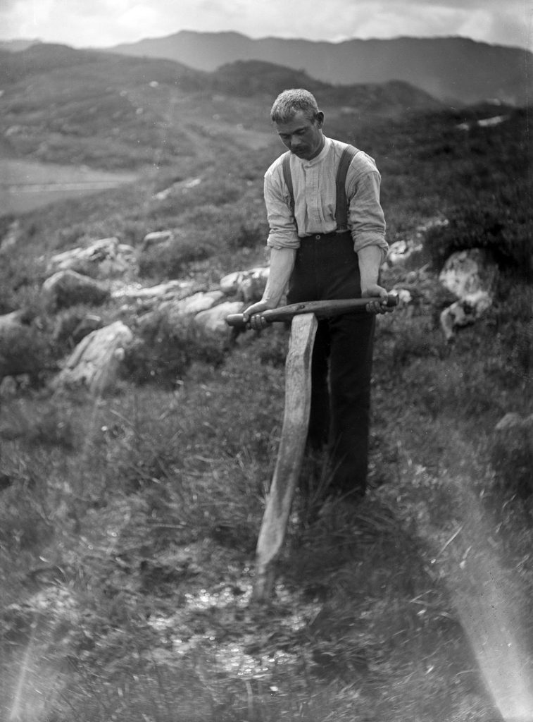 Black and white photograph of a man wearing a white shirt and trousers with suspenders, using both hands to work a wooden spade in a stony field. A range of hills extends in the background.