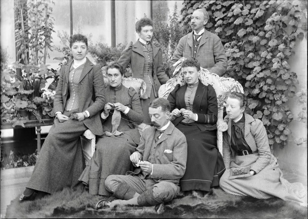Vintage black and white photograph of a group of two men and five women, all in formal attire, sitting in a glass conservatory surrounded by lush foliage.