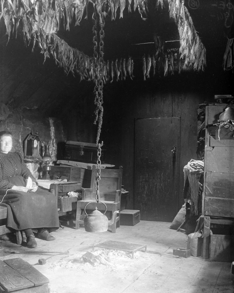 Old black and white image of a woman sitting by a central fire with a kettle hanging over it inside a traditional, austere stone and timber house.