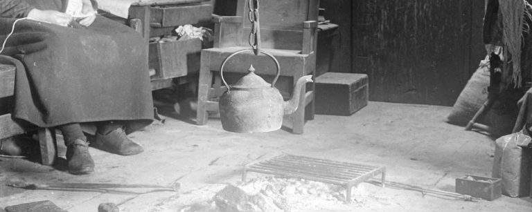 Black and white picture of the inside of an early 20th century home. A kettle hangs on a hook over a central fire and a woman knits in a chair on the left.