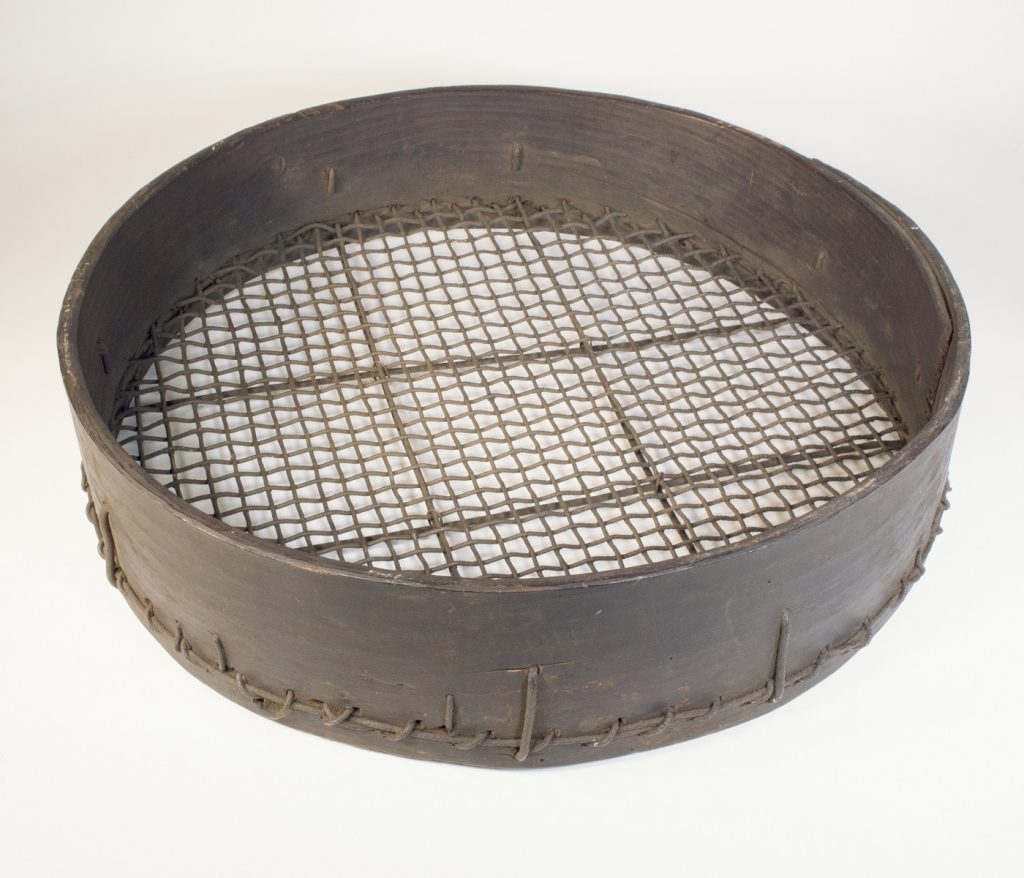 A brown mesh sieve with a thing, high wooden rim laying flat down on a cream-coloured surface. 