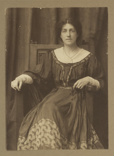 Sepia photo of May Morris sitting on a chair in a gown, with her arms resting on the chair's armrests and looking straight into the camera.