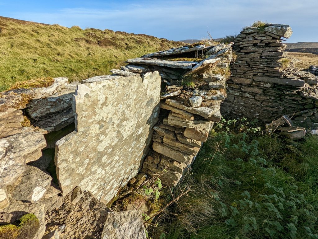Closeup of a section of wall within a roofless, ruinous cottage. A thin, upright, grey stone slab rises out from within a section of wall, identical to the stone slabs seen in the nearby ancient tombs.