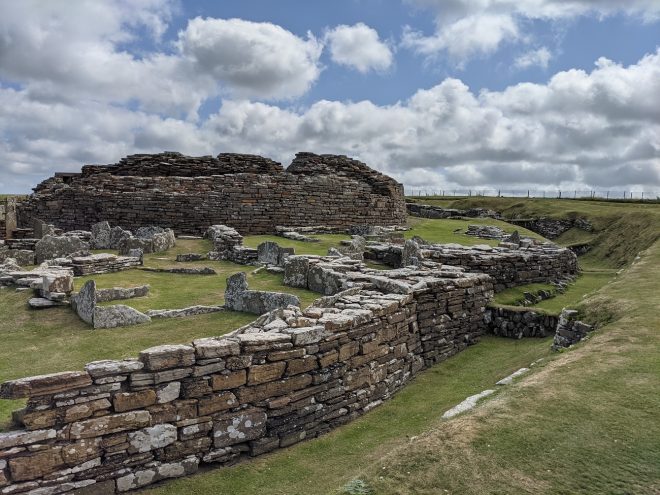 The circular remains of a stone tower surrounded by a scattering of stone slabs. To its right is a deep defensive, grassy ditch lined with grey stones. The sky is blue with rolling clouds.