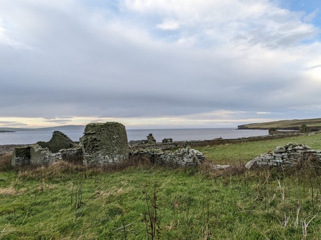 Light grey clouds and a blue sky linger over a scramble of grey stone ruins along a rugged shoreline. On the left a cylindrical tower rises to first floor level, and several low stone walls branch out from it. Headlands can be seen on both sides in the distance across the Eynhallow Sound.