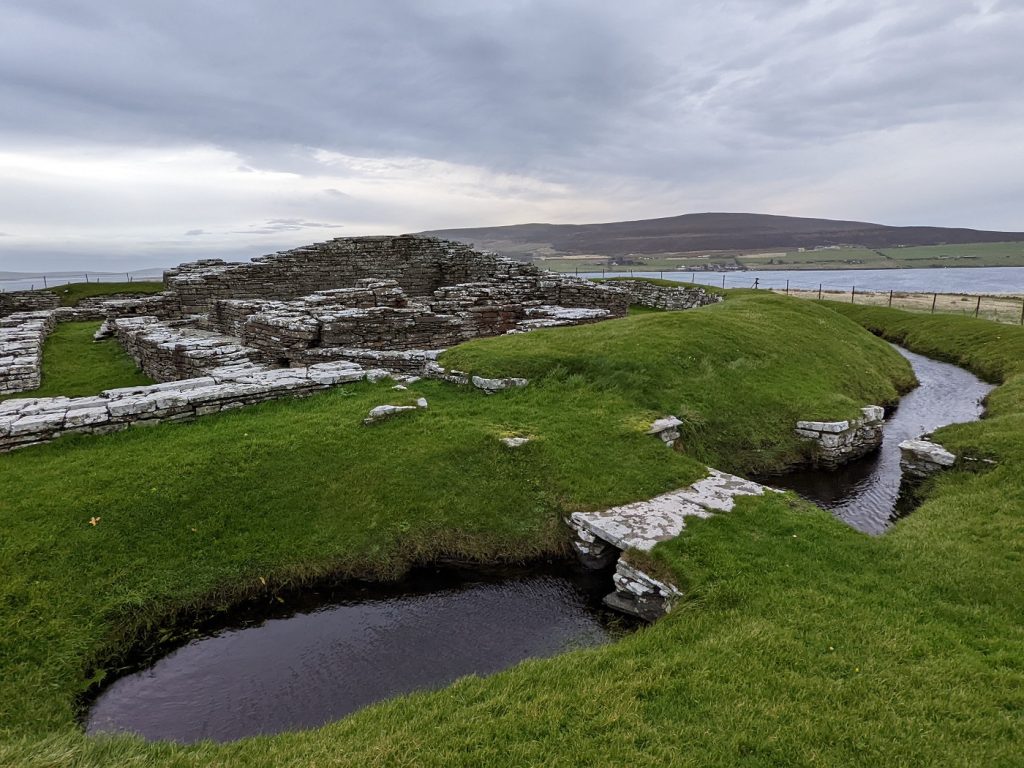 The ruins of a grey stone castle surrounded by a ditch partially filled with water. The bright green grass contrasts with the grey stone and grey sky. The castle ruins are squat and only rise to first floor level. A channel of water and the island of Rousay are in the distance.