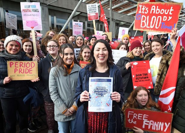 A demonstrating group of young women stand together holding signs advocating for free period products. In the centre, MSP Monica Lennon smiles and holds up a sign reading 'Period Products Free Provision Scotland Bill'. 