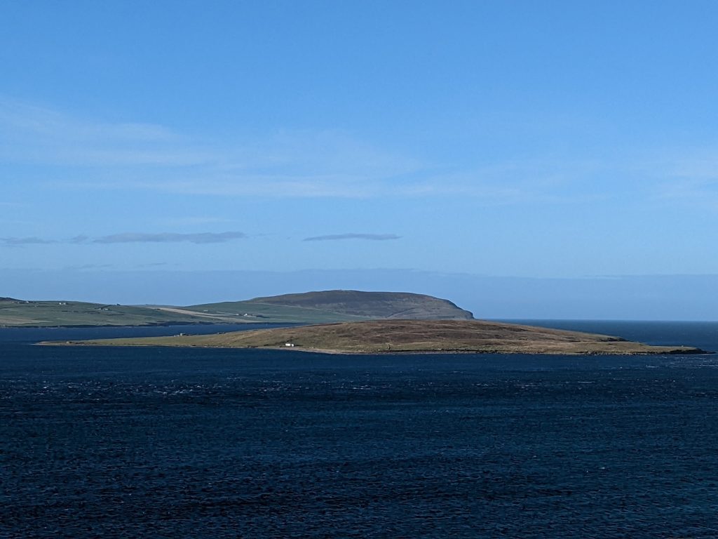 A wide landscape view over a deep blue channel of water. A low whaleback-shaped island is in focus in the middle, with green-brown grass and a single tiny building. Waters churn around the island, and a high cliff face rises in the distance.