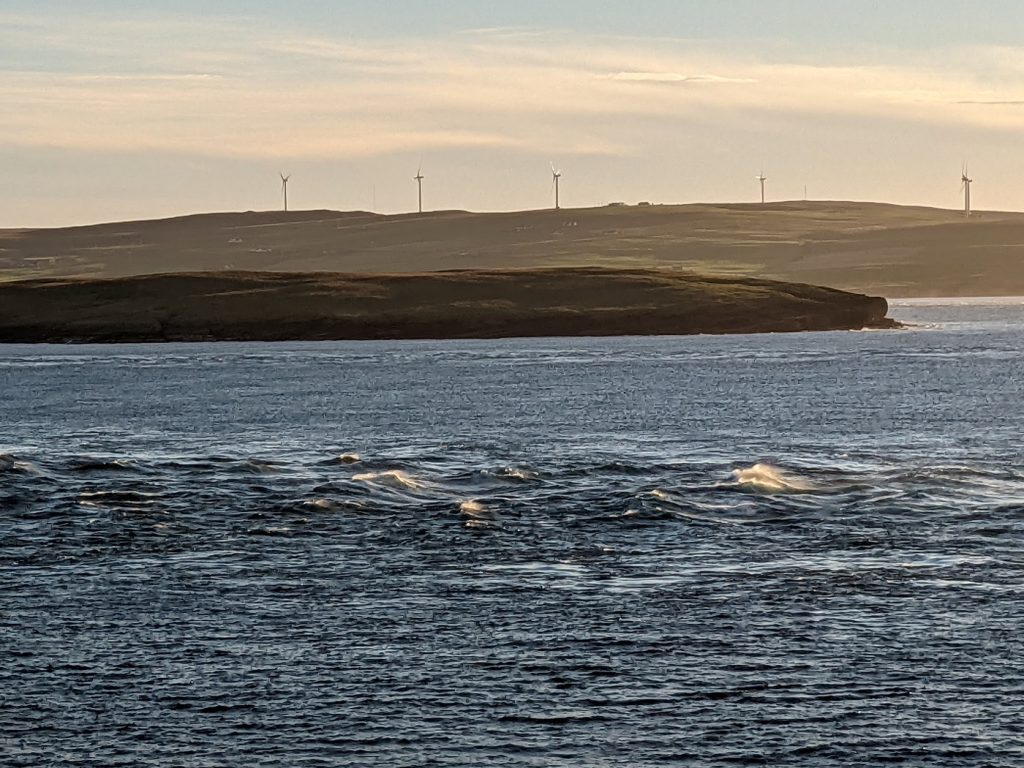 A soft yellow sunset over a channel of water, with an island and distant headland dominating the horizon. In the foreground, a strip of water in the channel is marked by sudden churning waves while the waters around it are fairly still.