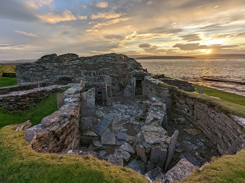 A breathtaking yellow sunset casts light on the massive stone ruins of Midhowe Broch. In the foreground, a sunken area of stone walls and partitions is enclosed by grassy banks. Beyond it, a ruinous round stone tower rises to first floor level.