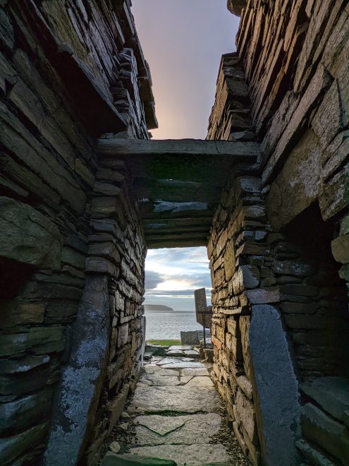 A monumental view from within the tall but narrow stone entrance passage of Midhowe Broch. Vertical stone walls flank the viewer with a flat stone slab capping the roofless entrance. Through the passage you can see a distant island over the water, with a cloudy purple sunset.